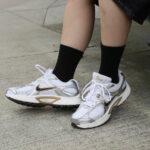 Person wearing white Nike sneakers with gold swoosh and black socks standing on a concrete sidewalk.