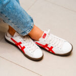 Pair of white sneakers with red side stripes on a tiled floor, jeans cuffs visible.
