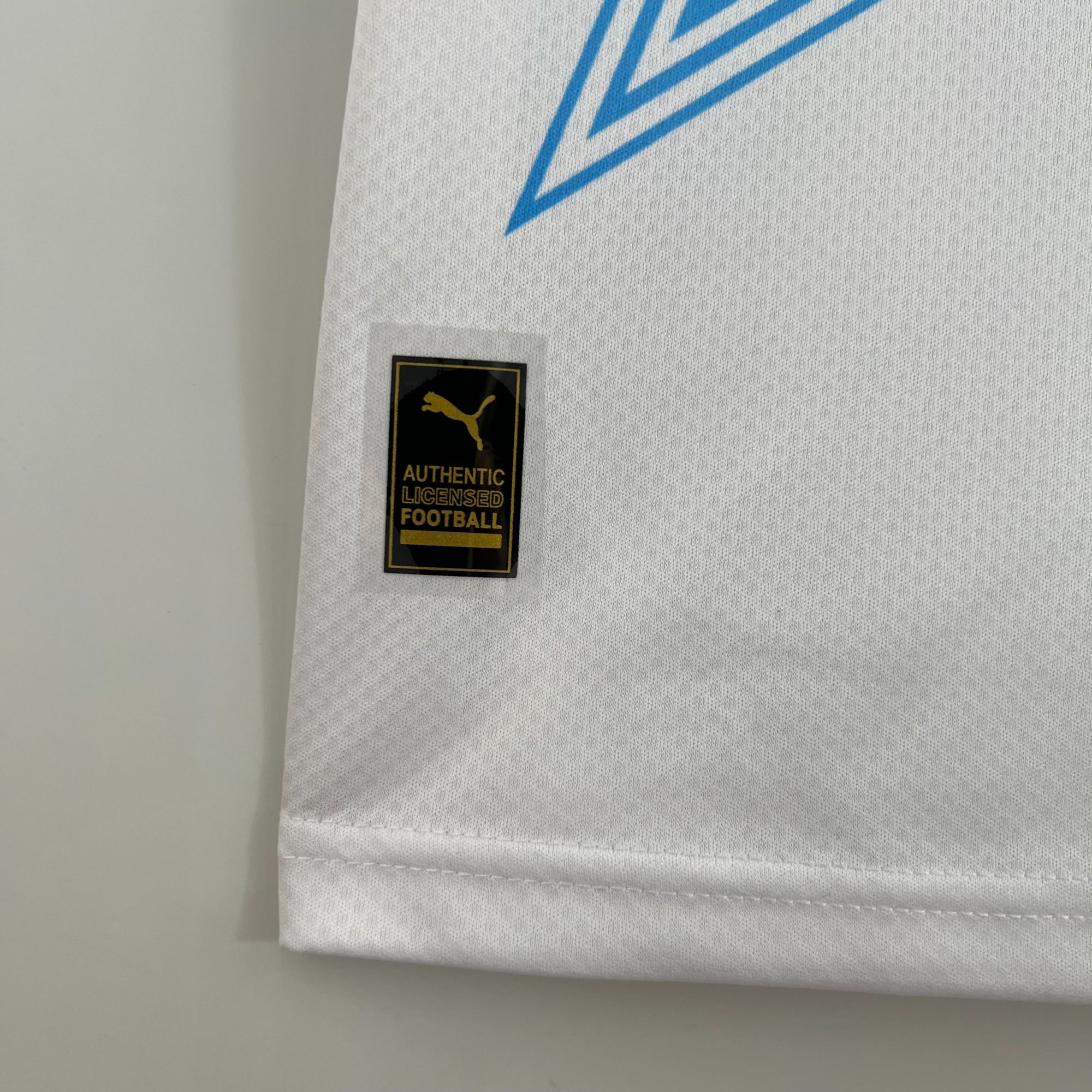 3c44edc6_aa10e4e0-58fd-4584-9599-c96028ef5ac6 | UAE SPORT STORE Close-up of white fabric with blue geometric print and a black-and-gold Authentic Licensed Football badge sewn onto the seam.