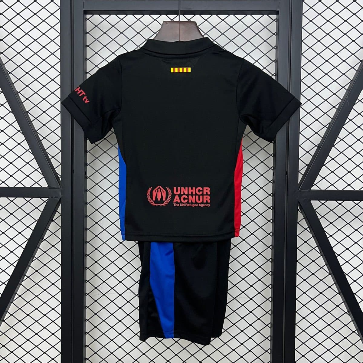 Back view of a black athletic jersey hanging on a wooden hanger, with blue and red side panels and the UNHCR logo and text near the bottom main back area, against a metal grid backdrop with a small yellow-red striped patch at the upper center.