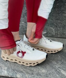 Person tying white athletic sneakers with a thick beige sole on a stone surface; red sleeves visible in the background.