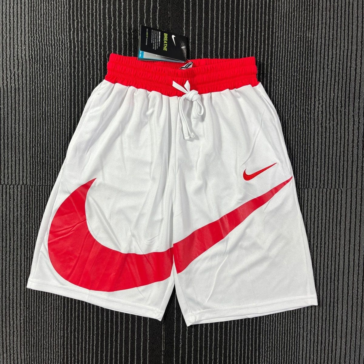 White athletic shorts with a red elastic waistband and red diagonal Nike swoosh marks on a dark textured carpet backdrop, tag visible on the waistband edge.
