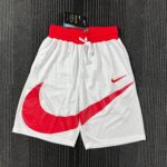 White athletic shorts with a red elastic waistband and red diagonal Nike swoosh marks on a dark textured carpet backdrop, tag visible on the waistband edge.