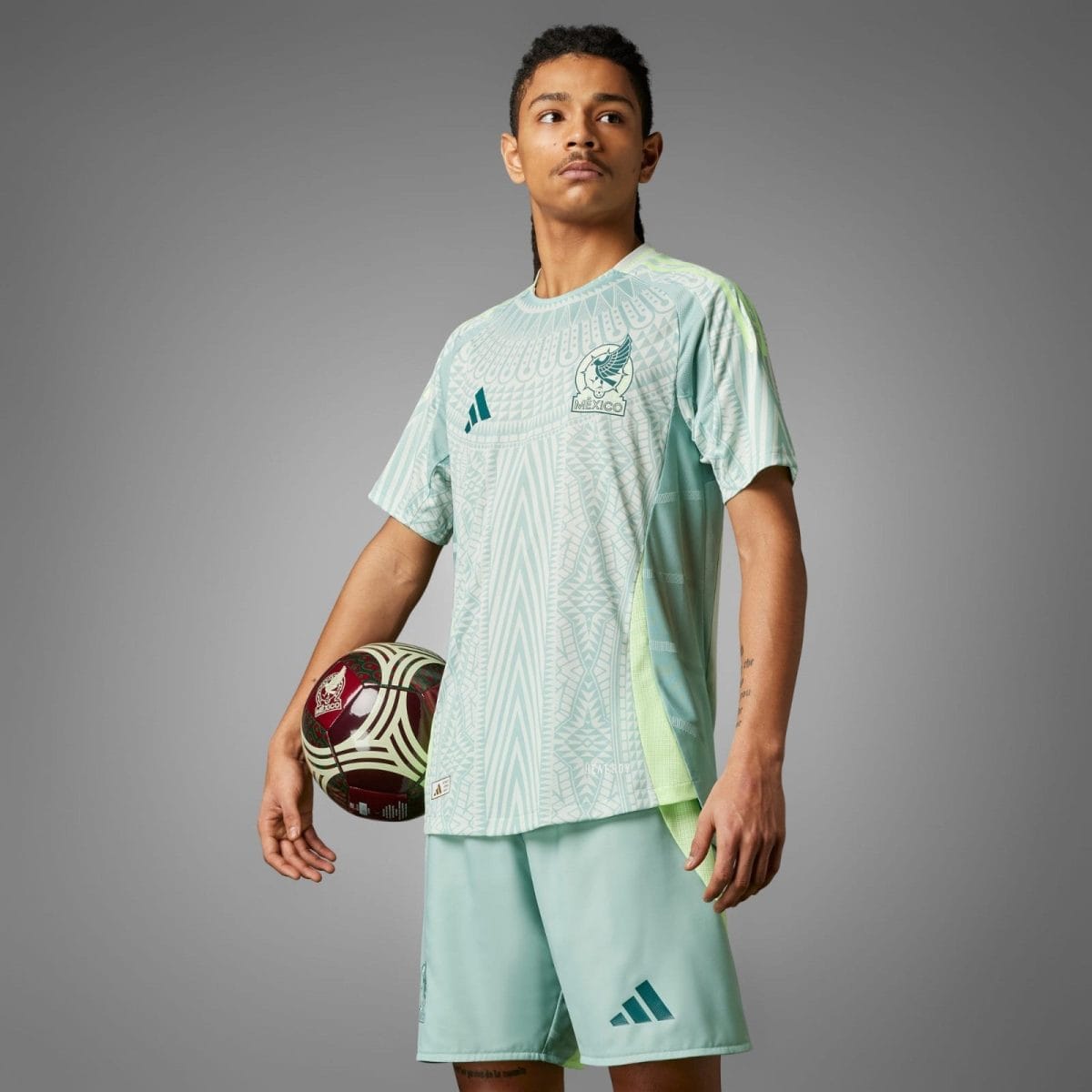 Young male soccer player in a light teal Mexico jersey holding a patterned soccer ball, standing against a gray background.