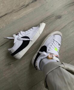 Pair of white sneakers with a black Nike swoosh on a light wooden floor; one shoelace is untied and loosened.