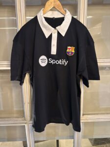 Black polo-style shirt on a hanger with a white collar, FC Barcelona crest, and Spotify logo on the chest.
