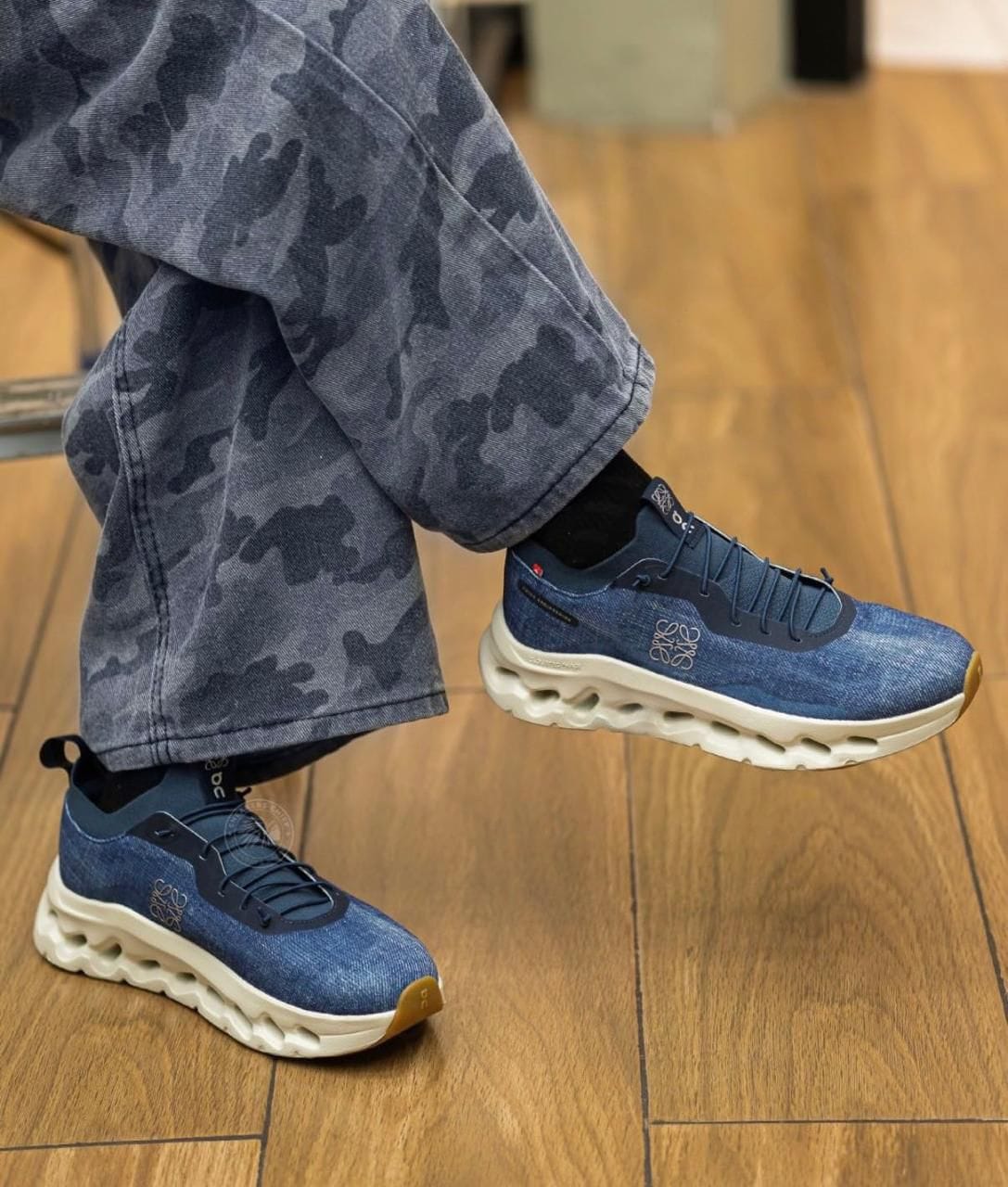 3e8e04d3-ad5f-4b6d-92b0-76ee4a8e755d | UAE SPORT STORE Blue denim-textured running sneakers with a chunky white sole, worn with gray camo jeans on a wooden floor.