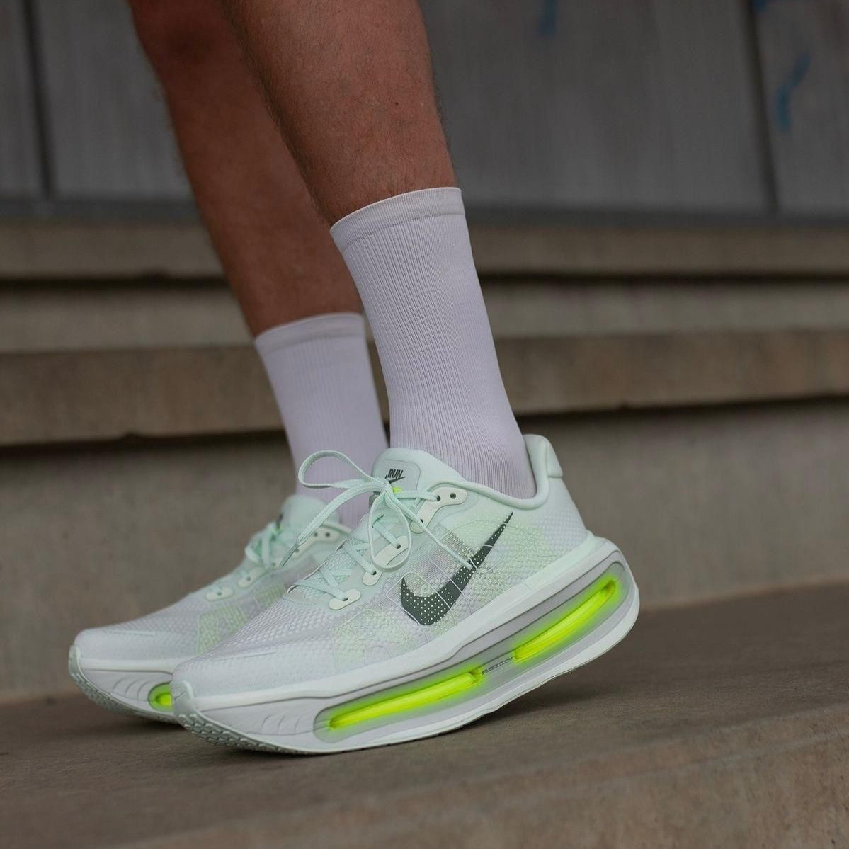 3f0995ba-67e9-453c-bd2f-2af14a249f76 | UAE SPORT STORE White Nike running shoes with neon green air-cushioned soles, worn with white socks on a concrete step backdrop.