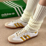 White Adidas sneakers with yellow stripes and cream suede toe caps, paired with thick ribbed cream socks, beside a green Adidas shoebox on carpeted floor.