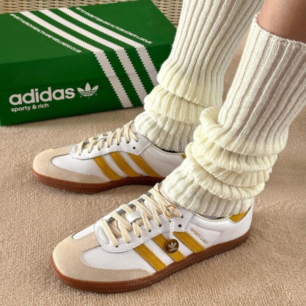 White Adidas sneakers with yellow stripes and cream suede toe caps, paired with thick ribbed cream socks, beside a green Adidas shoebox on carpeted floor.