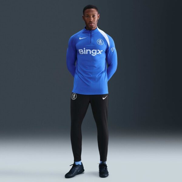 Male soccer player in a blue training top with crest and Bingx sponsor, black joggers, standing with hands behind back against a dark backdrop.