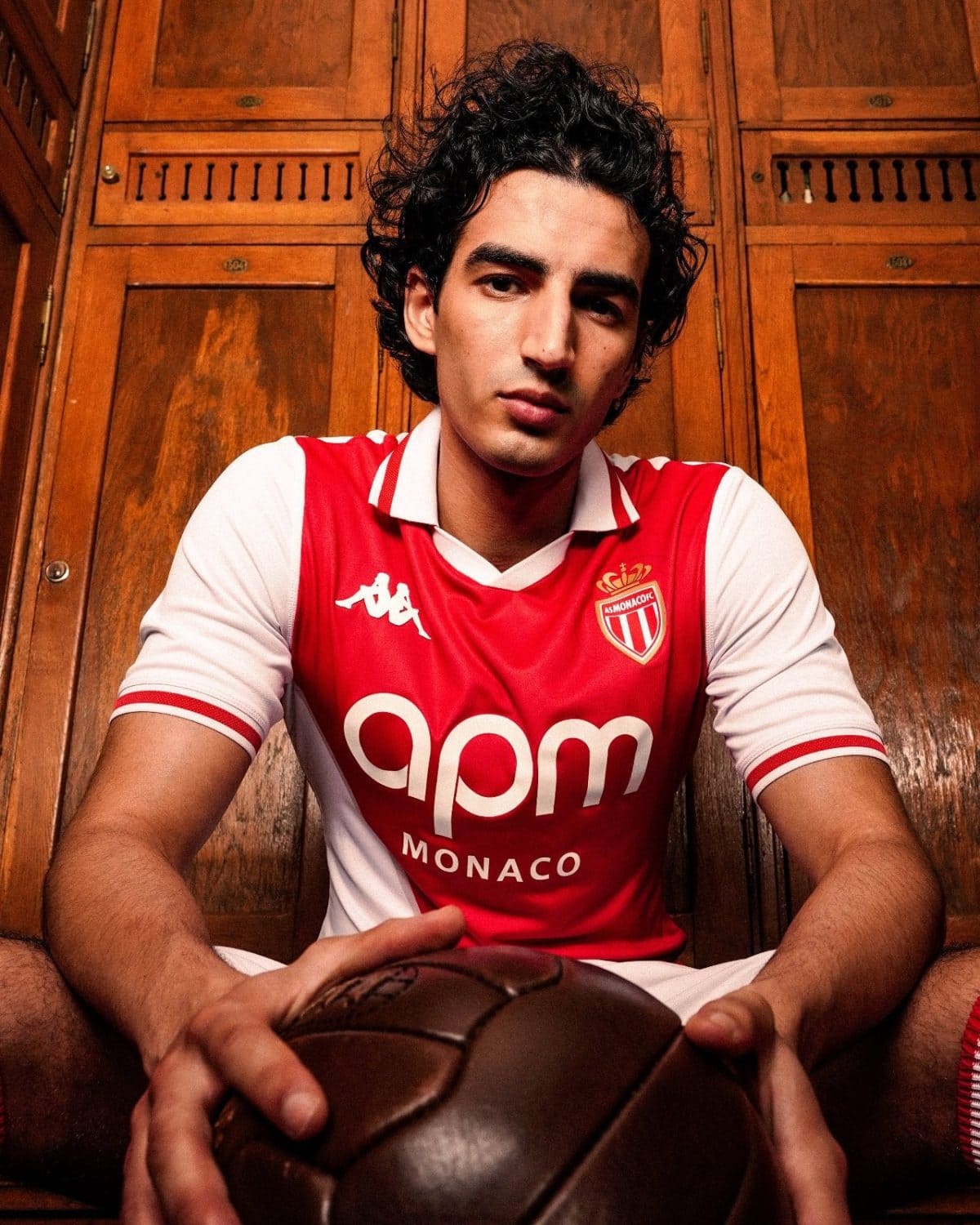 Young man in AS Monaco football jersey seated in a wooden locker room, holding a brown leather football toward the camera.