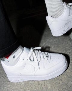 Two white Nike sneakers worn with white socks, with one foot on the ground and the other resting on a raised surface.