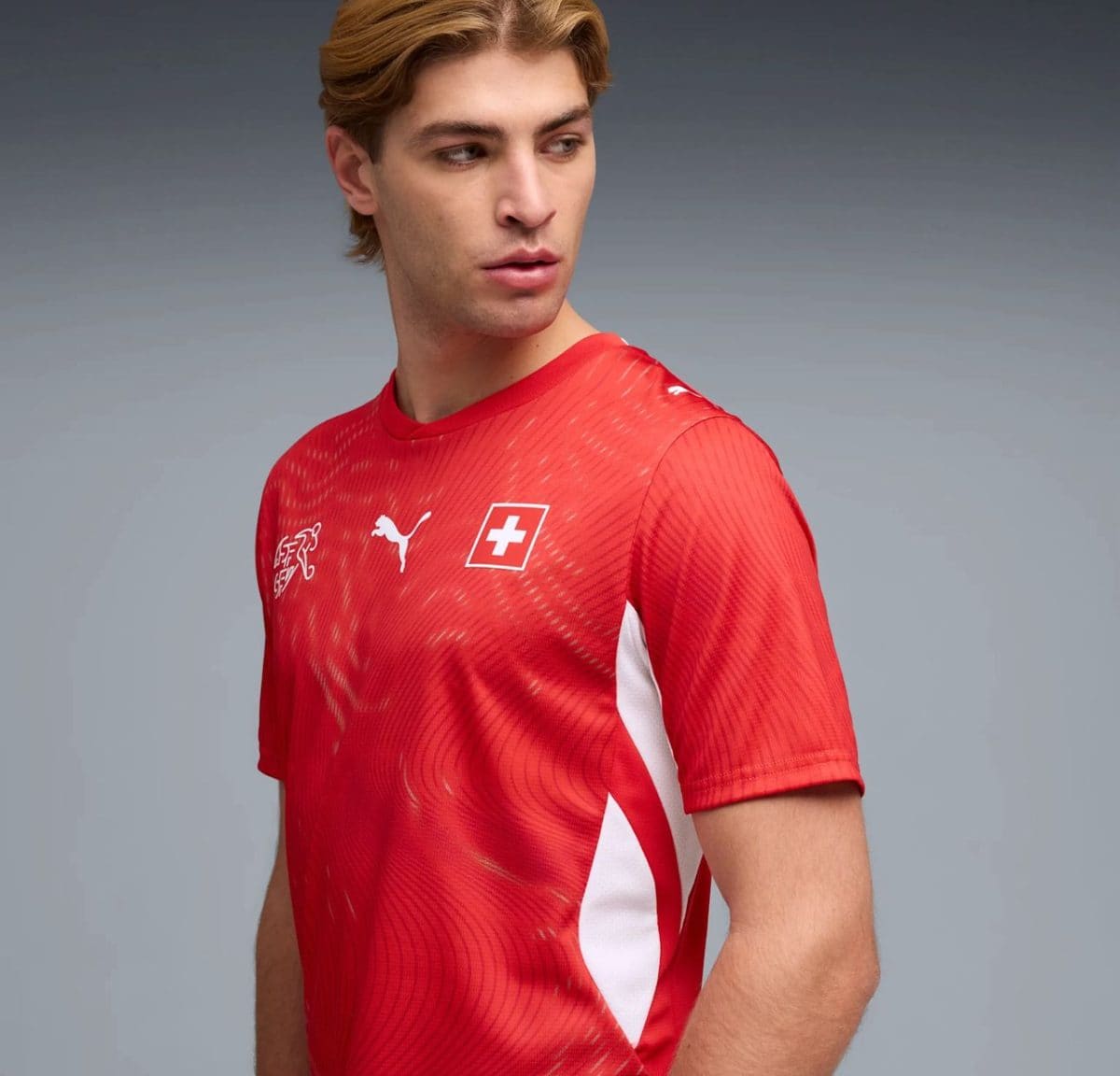 Male athlete in a red Swiss soccer jersey looking to the side, against a neutral gray background.