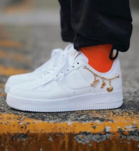 White Nike Air Force 1 sneaker with bright orange sock and a gold charm bracelet around the laces on a curb, front view.