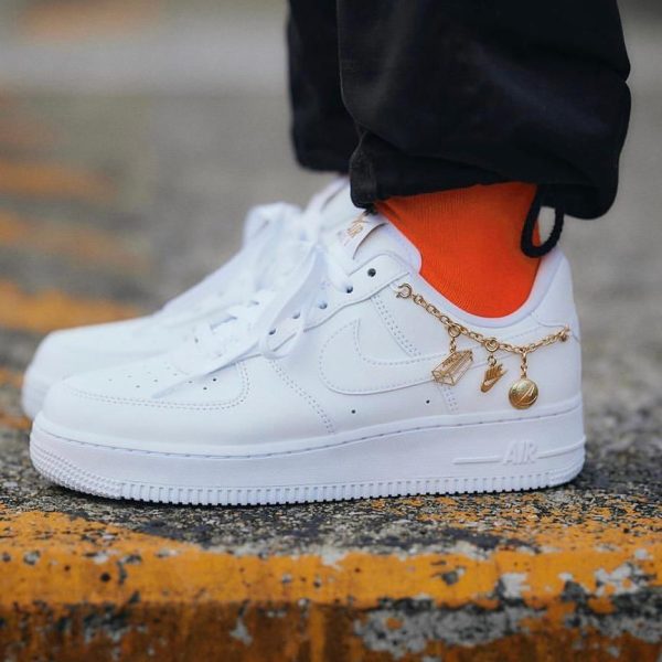 White Nike Air Force 1 sneaker with bright orange sock and a gold charm bracelet around the laces on a curb, front view.