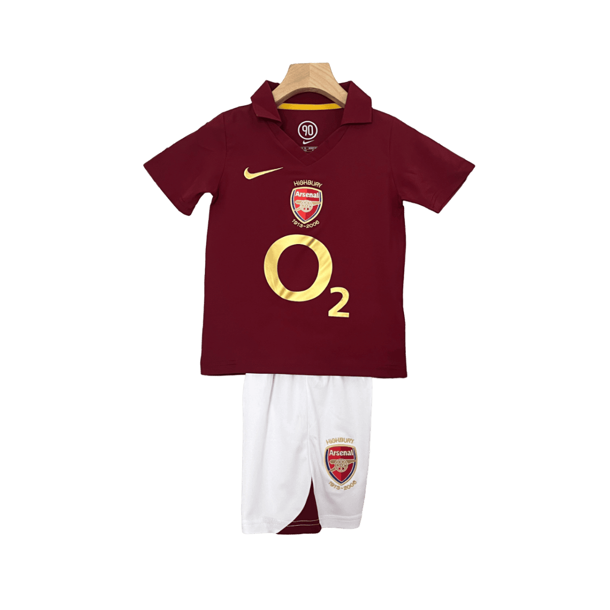 Maroon Arsenal football jersey with gold O2 logo on a hanger, paired with white shorts featuring the club crest.