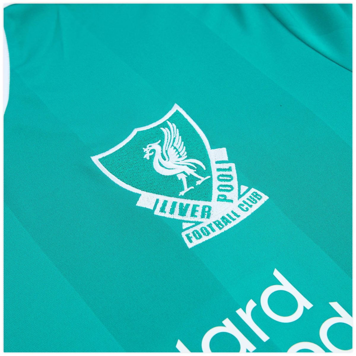 Teal fabric with a white embroidered shield logo and partial text, likely a sports club crest. The design is diagonally cropped across the image.