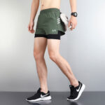 40810641 | UAE SPORT STORE Man wearing olive green athletic shorts over black compression shorts, black Nike running shoes, and a wristwatch while holding a towel at his side.