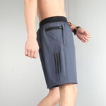 Side view of a man wearing gray-blue athletic shorts with a zippered rear/side pocket and three black stripes on the leg, black waistband showing beneath a bare torso.