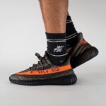 Foot wearing a patterned Yeezy Boost 350 sneaker with an orange SPLY-350 stripe and thick black sole, paired with black striped socks.