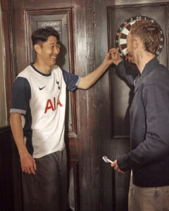Two men celebrate with a high-five beside a dartboard in a wooden-bar setting; one wears a Tottenham jersey, the other holds a dart.