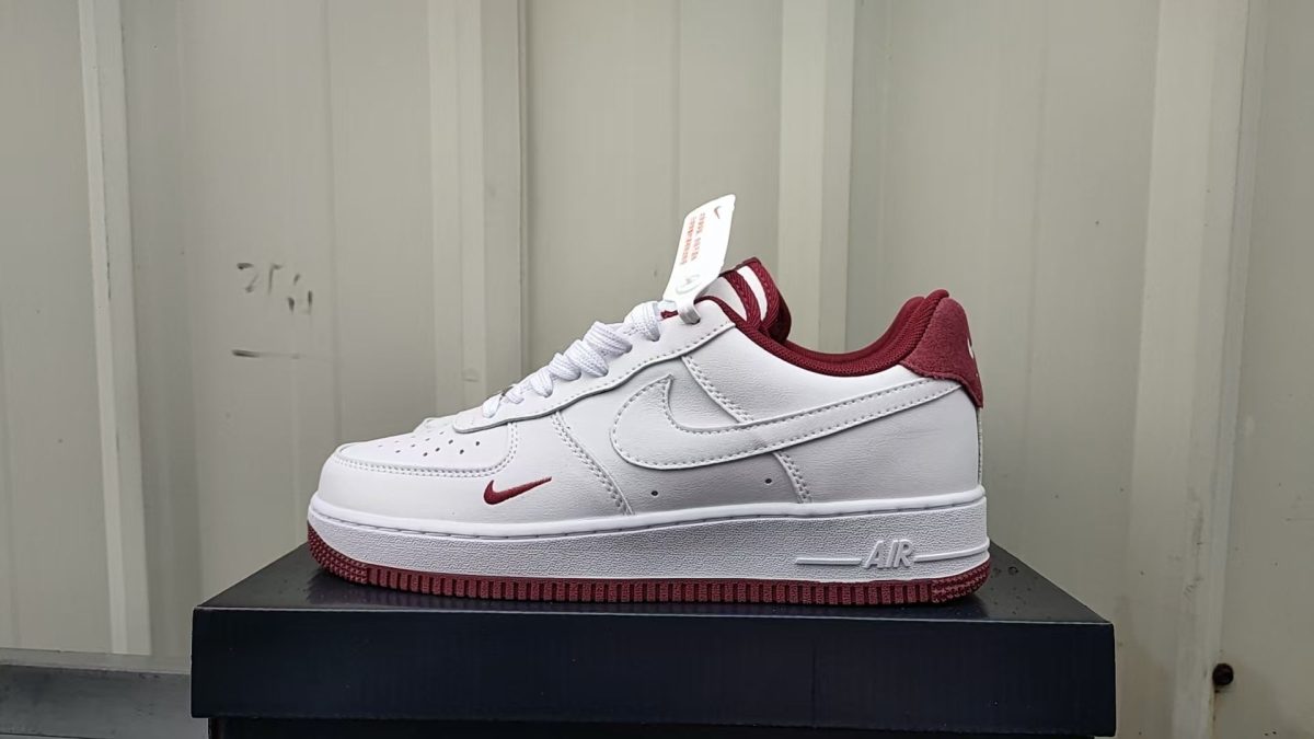 White Nike Air Force 1 sneaker with maroon accents sits on a black box; visible swoosh, maroon heel, and tag on the tongue.