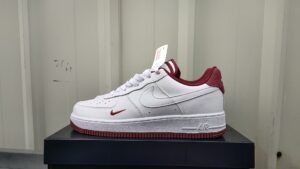 White Nike Air Force 1 sneaker with maroon accents sits on a black box; visible swoosh, maroon heel, and tag on the tongue.