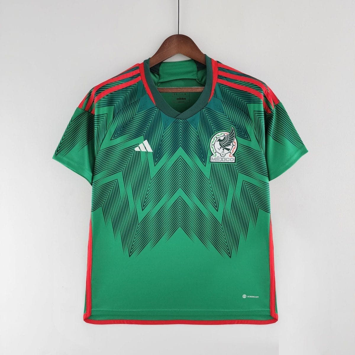 Green soccer jersey on a wooden hanger with red trim and bold geometric pattern, featuring a Mexico crest and Adidas logo near the chest area.