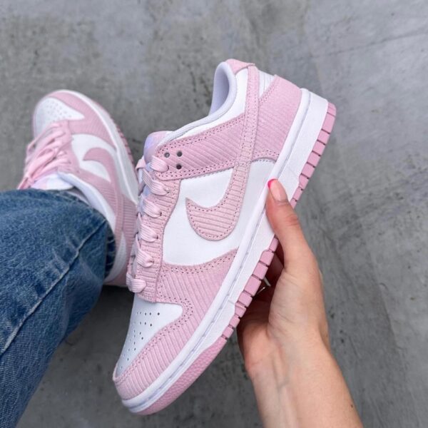 Pink and white Nike sneaker being held by a hand, denim jeans in the background on a gray concrete floor.