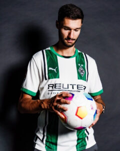 Male soccer player in a white and green jersey holds a colorful soccer ball, looking down at it against a dark backdrop.