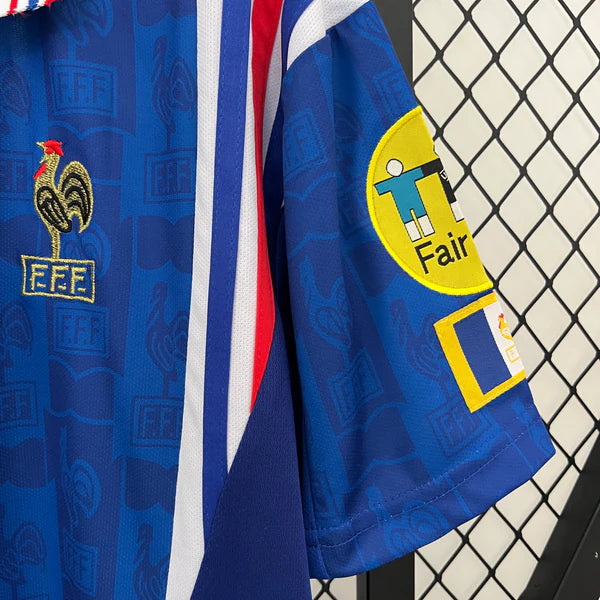 41d74386 | UAE SPORT STORE Close-up of a blue sports jersey with FFF crest and a yellow 'Fair' patch on the sleeve, against a chain-link fence.