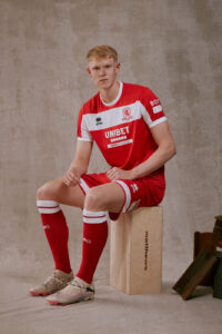 Young male soccer player in red-and-white kit sits on a wooden box against a neutral backdrop.