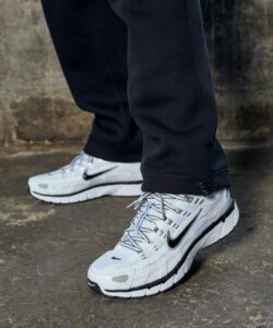 White Nike running shoes on a person wearing black pants, standing on a concrete surface.