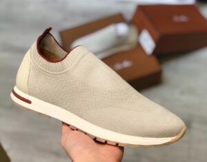 42b24849-bed6-4c52-be17-ea2d0acecc6d | UAE SPORT STORE Beige knitted slip-on sneaker with a white sole, held in hand with blurred brown shoe boxes in the background.