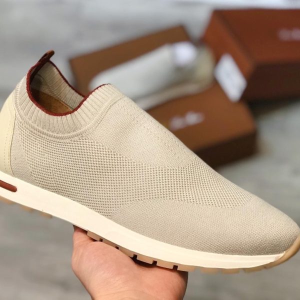Beige knitted slip-on sneaker with a white sole, held in hand with blurred brown shoe boxes in the background.