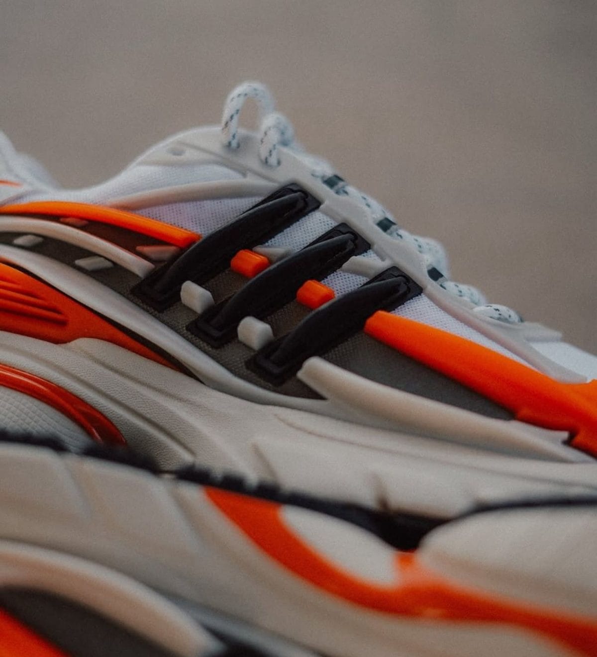 White athletic sneaker with orange accents and black lace eyelets viewed from the side, showing layered sole details.
