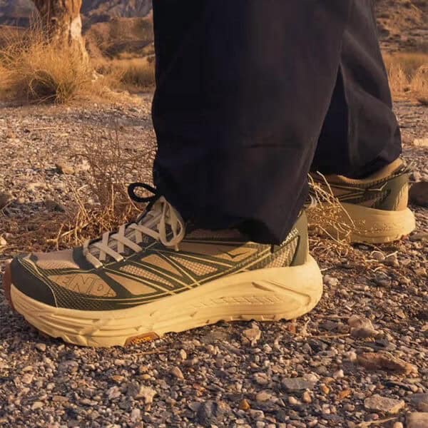 Pair of green and beige trail running shoes on a rocky desert surface with black pants cuffs visible.