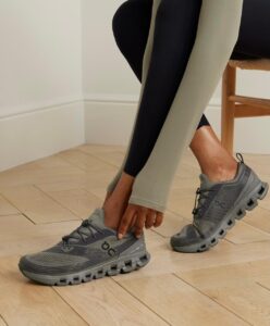 Person tying gray athletic sneakers with a chunky segmented sole while wearing olive leggings.