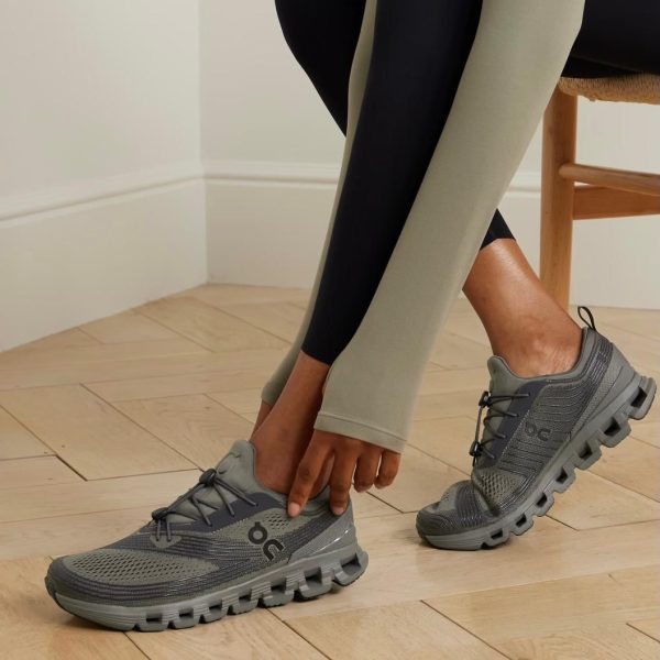 Person tying gray athletic sneakers with a chunky segmented sole while wearing olive leggings.