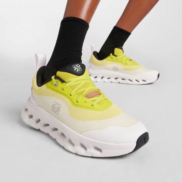Bright yellow chunky sneakers with white thick sole on a model wearing black socks, close-up view of one foot in front of the other on a gray background