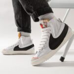 White high-top sneakers with a black side panel, worn with cuffed jeans in a bright studio setting.