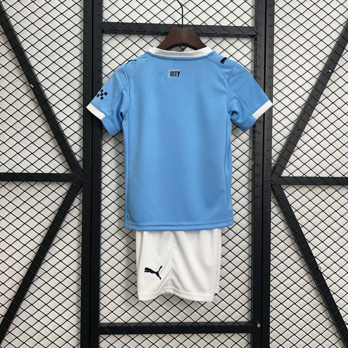 Light blue sports jersey on a wooden hanger, seen from the back, with a white collar and the word CITY on the upper back. A white garment with a dark Puma logo hangs below against a black metal grid wall.