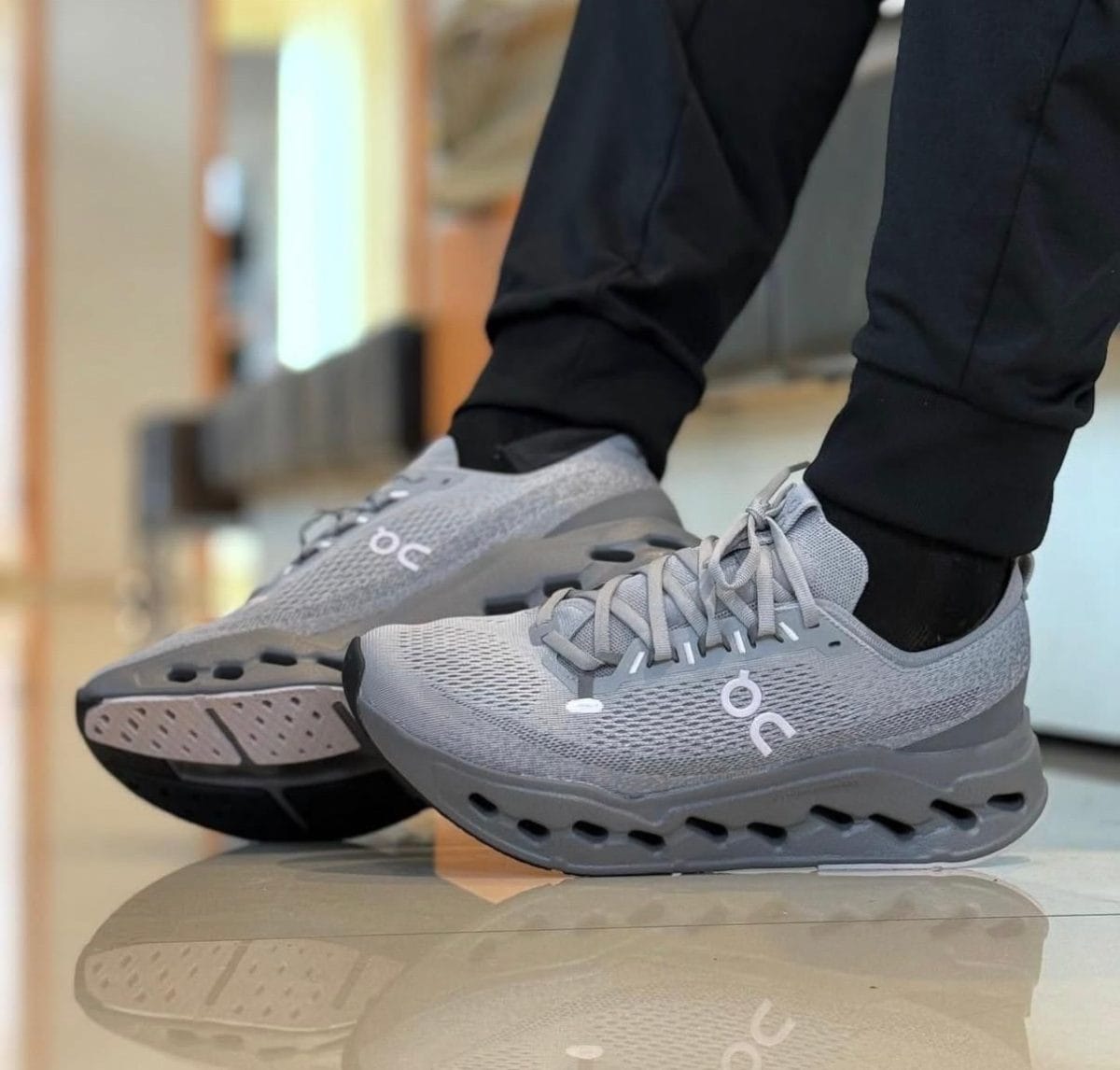 Gray On running shoes with perforated mesh, laces tied, worn by someone in black pants standing on a glossy floor.