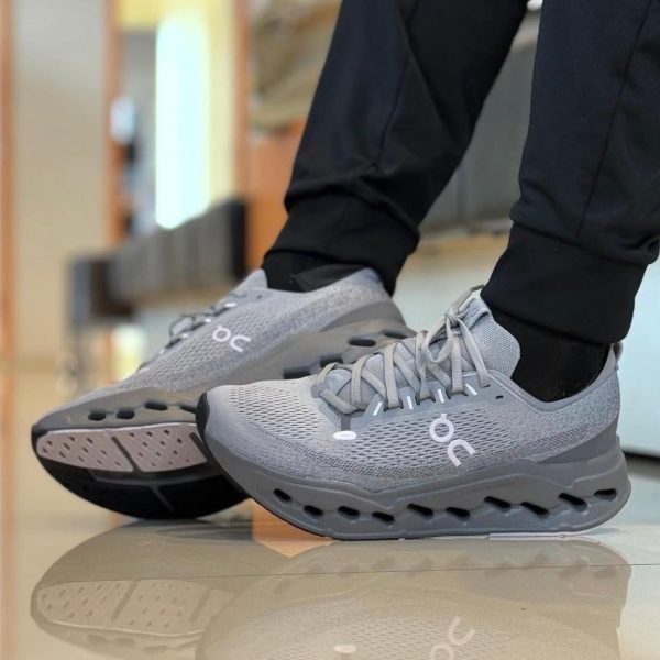 Gray On running shoes with perforated mesh, laces tied, worn by someone in black pants standing on a glossy floor.