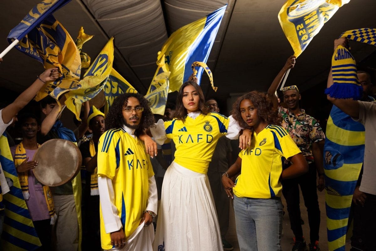 Group of fans in yellow and blue jerseys celebrate, waving flags at a crowd-filled stadium.