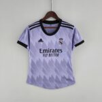 Light purple Real Madrid jersey on a wooden hanger against a pale wall, displaying Emirates Fly Better text and Adidas logos.