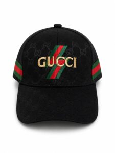 Black baseball cap with Gucci monogram pattern and a gold-embroidered Gucci logo over a green–red stripe motif on the front.