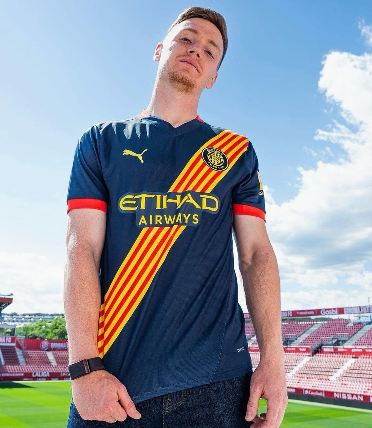 Young man wearing a blue Girona FC jersey with red-yellow diagonal stripe, standing in a stadium against a blue sky.