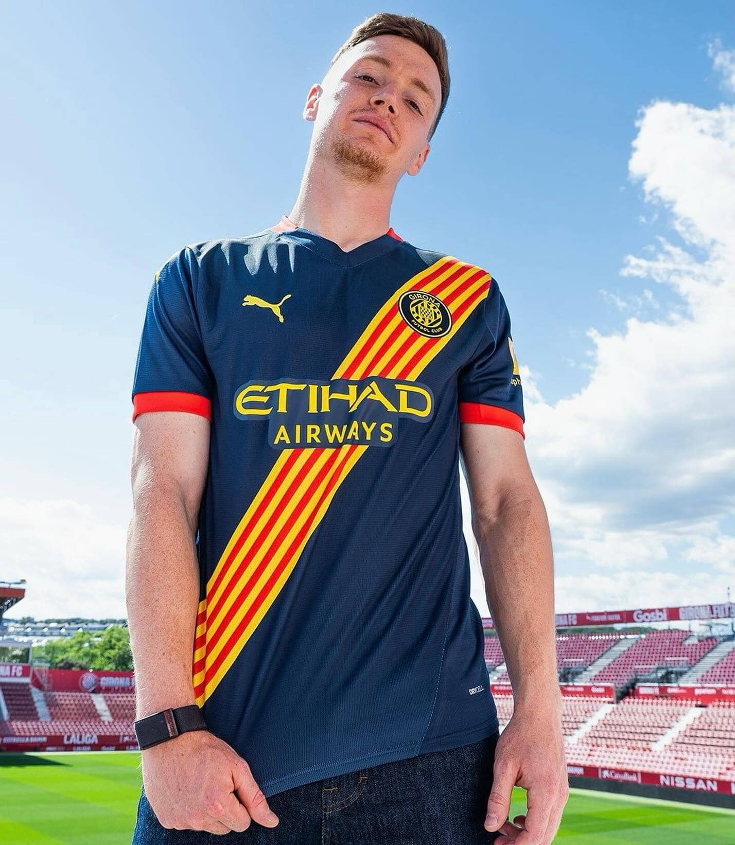 461505398_17940808712903197_5106101443471822670_n | UAE SPORT STORE Young man wearing a blue Girona FC jersey with red-yellow diagonal stripe, standing in a stadium against a blue sky.